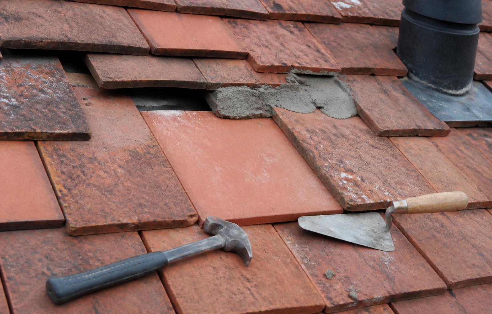 Close-up of a pitched clay tile roof mid-repair with a freshly bedded replacement tile, fresh mortar and hand tools resting on the tiles